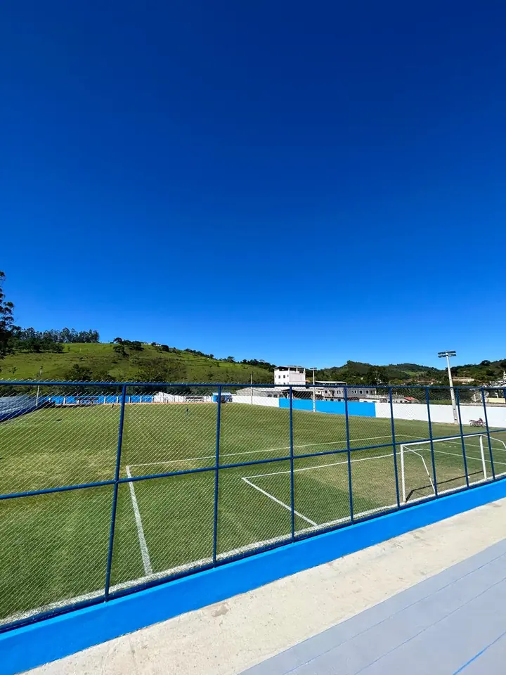 Campo de futebol reformado em Mercês, com gramado, alambrado azul e muretas pintadas, sob céu azul